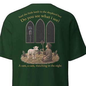 Puede incluir: Una camiseta verde oscuro con un gráfico de un joven pastor y un cordero arrodillados sobre heno. Arriba hay tres ventanas arqueadas y el texto: Said the little lamb to the shepherd boy Do you see what i see? A cam, a cam, watching in the night.
