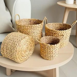 Peut inclure: Un ensemble de quatre paniers ronds en jonc de mer naturel tressé de couleur brun clair. Chaque panier présente une texture épaisse et tressée avec deux petites anses latérales robustes. Les paniers sont de tailles variées et groupés.