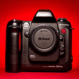 Puede incluir: Una cámara digital Fujifilm FinePix S2 Pro de color negro con montura de objetivo Nikon F. El cuerpo de la cámara muestra el logotipo FinePix en la parte superior, la marca Fujifilm y la designación del modelo S2 Pro en el panel frontal.