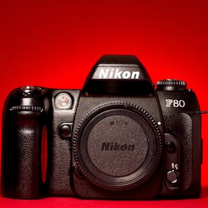 Op de afbeelding: Een zwarte Nikon F80 35mm filmcamerabehuizing met een getextureerde grip. De voorzijde toont het Nikon-logo in wit, de modelnaam F80 en een ronde lensvattingdop met het woord OPEN en het Nikon-logo in reliëf in het midden van het zwarte plastic.