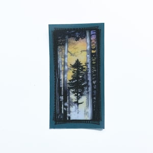 Könnte beinhalten: Ein blaugrüner Stoff-Patch mit einem schwarzen Umriss eines Kiefernbaums in einer Waldlandschaft. Der Hintergrund ist ein aquarellartiger Sonnenuntergang in Gelb-, Orange-, Lila- und Blautönen.