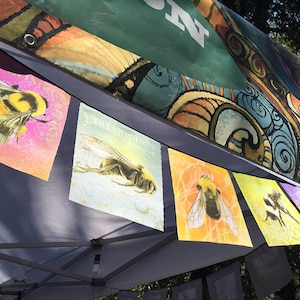 May include: Five colorful fabric pennants with illustrations of bumblebees. The pennants are hanging from a white awning. The pennants are decorated with floral patterns and have a vintage aesthetic.