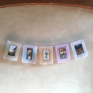 May include: A string of five colorful fabric pennants with a tree design and text. The pennants are hung on a string against a light beige wall. The text on the pennants reads "We All Are Free Let Us Be Free".