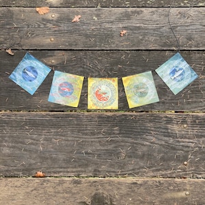 May include: A string of five colorful flags hanging from a string. Each flag has a different design featuring a fish, a flower, and other nature-inspired elements.