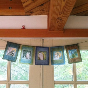 May include: A string of six colorful fabric pennants with owl illustrations hangs in front of a window. Each pennant features a different owl design with a blue, green, and brown color scheme.