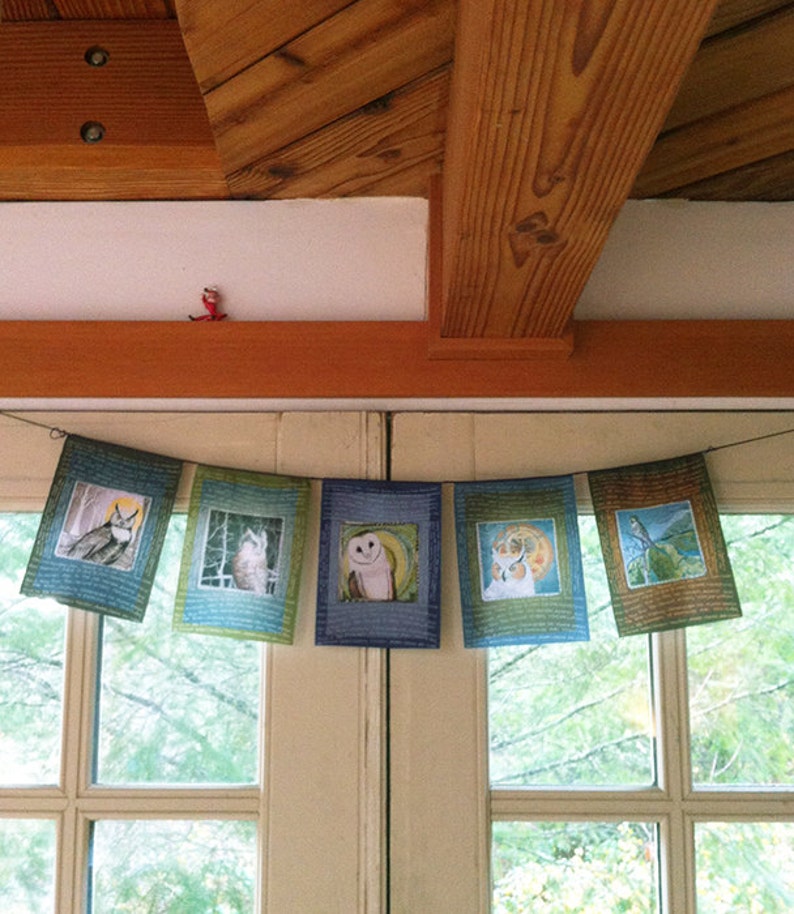 May include: A string of six colorful fabric pennants with owl illustrations hangs in front of a window. Each pennant features a different owl design with a blue, green, and brown color scheme.