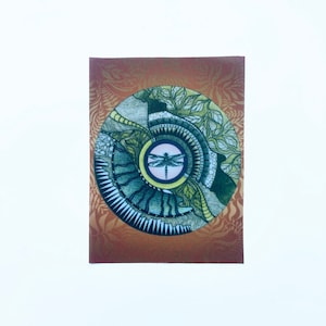 May include: A circular mandala design with a dragonfly in the center. The mandala is made up of intricate patterns in shades of green, brown, and white. The background is a warm orange color.