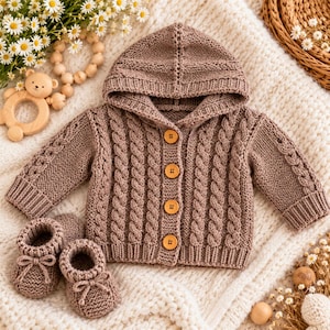 Può includere: Un cardigan in maglia color marrone chiaro con cappuccio, caratterizzato da un motivo a trecce verticali e quattro bottoni in legno sul davanti, accompagnato da un paio di scarpine in maglia abbinate con piccoli fiocchi sul davanti.