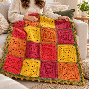 May include: A handmade crochet blanket featuring a grid of square motifs in vibrant shades of yellow, orange, and magenta. The squares are joined with an olive green yarn, and the entire blanket is finished with a matching olive green scalloped border.