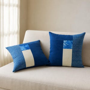 May include: Two square throw pillows featuring a patchwork design of deep blue solid fabric, a textured blue grid pattern, a light cream rectangular panel, and a small bright blue marbled accent square, arranged on a light-colored sofa.