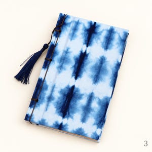 May include: A rectangular journal featuring a blue and white tie-dye pattern on the cover. The binding is secured with dark brown cord and includes a matching dark blue decorative tassel attached to the spine.