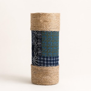 May include: A cylindrical vase wrapped in natural tan jute twine at the top and bottom sections. The center features a patchwork of dark blue fabric panels with various white and olive green geometric, floral, and grid patterns stitched together.