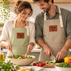 Pode incluir: Dois aventais listrados com remendos retangulares de couro marrom. Um avental é verde sálvia com o nome Emma, e o outro é bege com o nome Noah e um ícone de colheres. Ambos possuem alças de pescoço ajustáveis na cor creme.
