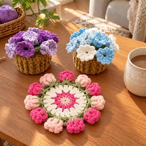 May include: Two crocheted potted flower arrangements in brown textured pots, one with purple and lavender blooms and one with blue and white blooms, alongside a circular crocheted coaster featuring a pink, white, and green floral pattern.