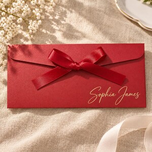 May include: A rectangular red envelope featuring a centred red satin ribbon tied in a bow. The name Sophia James is printed in gold cursive script on the lower right side of the envelope front, which has a smooth, matte finish.