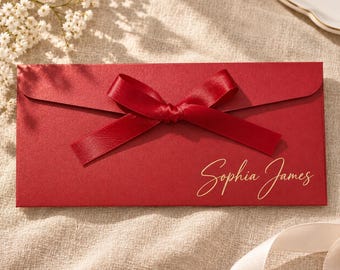Puede incluir: Un sobre rectangular de color rojo con un lazo de satén rojo atado en el centro. El nombre Sophia James está impreso en letra cursiva dorada en la parte inferior derecha del frente del sobre, que tiene un acabado mate y suave.