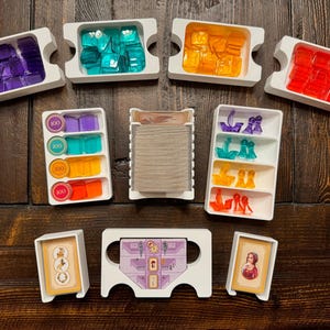 May include: A board game organiser set featuring white plastic trays containing translucent purple, teal, orange, and red square tokens, ship-shaped game pieces, circular 100-value markers, a tall deck of cards, and two individual card holders.