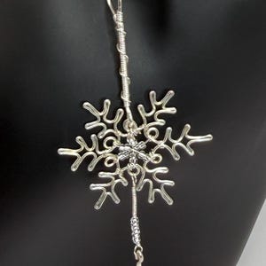 May include: A silver-toned wire-wrapped snowflake pendant suspended from a matching silver-toned chain. The snowflake design features intricate, hand-formed metal loops and coils, with a vertical wire extension ending in a small metal drop.