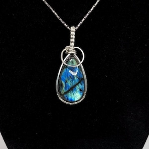 May include: A teardrop-shaped labradorite pendant featuring iridescent blue and black tones, encased in a silver wire-wrapped setting. The pendant hangs from a thin silver chain, showcasing a polished stone with natural dark inclusions.