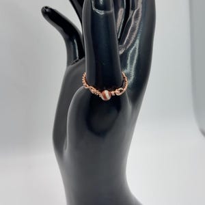 May include: A ring crafted from twisted copper wire featuring a single small, round, striped bead in shades of brown and white. The band is constructed from thin, metallic copper wire that is coiled and wrapped to secure the central bead.