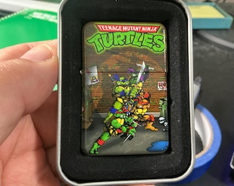 May include: A rectangular metal lighter featuring a graphic of the Teenage Mutant Ninja Turtles in a brick alleyway setting. The top of the lighter displays the Teenage Mutant Ninja Turtles logo in green and red text. The lighter is housed in a metal case.