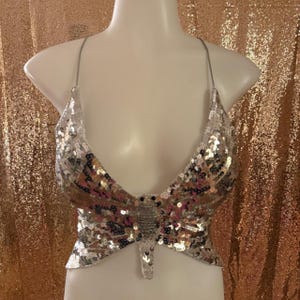 May include: A silver sequinned halter top shaped like a butterfly with thin straps. The garment features a central body section with wings extending outward, covered in reflective silver sequins that create a shimmering, textured surface appearance.