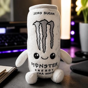 May include: A white plush toy shaped like a Monster Energy Ultra White can. It features the silver claw logo, the text ZERO SUGAR, and the text MONSTER ENERGY ULTRA WHITE. The plush has two small arms, two small legs, and a cute embroidered face.