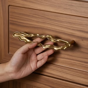 May include: A gold-toned metal drawer pull with an irregular, wavy, and organic shape. The handle features a textured, undulating surface that creates an open, elongated loop design, mounted horizontally on a flat, light brown wooden drawer front.