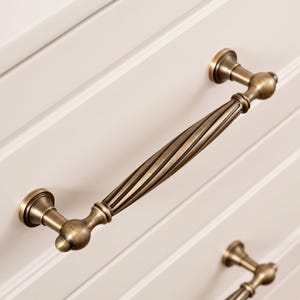 May include: A brass-toned metal drawer handle featuring a twisted, fluted centre bar with rounded ends and circular mounting bases. The hardware has a brushed finish with dark accents in the grooves, attached to a white panelled surface.