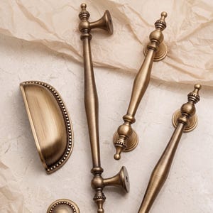 May include: A collection of antique brass cabinet hardware including one semi-circular cup pull with beaded edges, two long vertical bar pulls with decorative finials, and one small round knob, all featuring a brushed metallic finish.