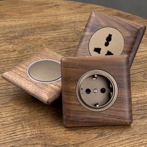 Può includere: Tre placche elettriche quadrate con finitura a venature di legno marrone scuro. Una placca presenta un interruttore circolare con un centro in bronzo metallizzato, mentre le altre due contengono prese elettriche circolari in una tonalità bronzo.