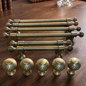 May include: A collection of antique brass cabinet hardware including four long bar pulls with spherical finials and five matching round knobs. Each piece features a textured, spiraled design on the ends and a smooth, brushed metallic finish.