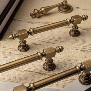 May include: A collection of antique brass-finished metal cabinet pulls and a single knob. The hardware features a cylindrical bar design with square mounting blocks, decorative finials, and circular bases with a brushed, weathered texture.