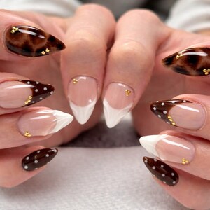 May include: Almond-shaped nails featuring a mix of designs including glossy dark brown tortoise shell patterns, white French tips, and dark brown tips with white polka dots, all accented with small gold metallic beads placed near the cuticles.