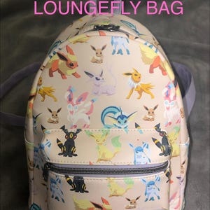 May include: A cream-colored mini backpack featuring an all-over print of various Eevee evolution characters. The bag has a front zippered pocket, a top handle, and a small rectangular brand patch at the bottom. Text at the top reads POKEMON LOUNGEFLY BAG.