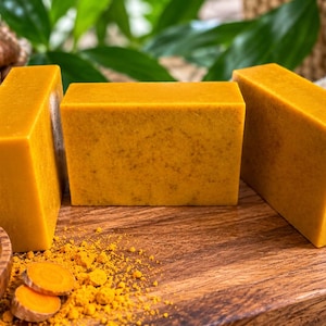 May include: Three rectangular bars of golden-yellow turmeric soap arranged upright. The soap has a smooth, solid texture with subtle flecks. Beside the bars are fresh turmeric roots, a small wooden bowl filled with turmeric powder, and slices.