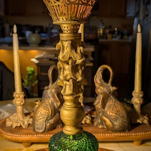 May include: A vintage brass table lamp with a green glass base and a decorative filigree shade. The lamp features a detailed, ornate design and is placed on a table with other decorative items, including candles and elephant figurines.