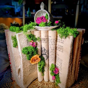 May include: A decorative open book with pages dusted in a glittery texture, adorned with bright green preserved moss and small dried pink and orange flowers. A small white ceramic teacup sits on top, filled with pink flowers and green moss.