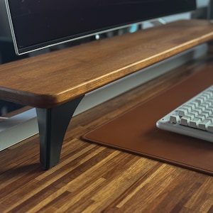 Op de afbeelding: Een houten monitorstandaard met een glad, rechthoekig blad en zwarte metalen poten. De standaard staat op een houten bureauoppervlak, voorzien van een bruine rechthoekige bureaumat en een lichtgrijs mechanisch toetsenbord ervoor.