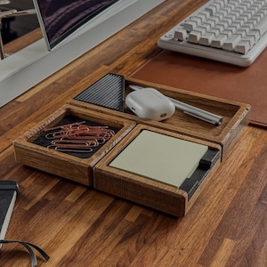 May include: A wooden desk organizer tray with three compartments. One section holds copper-colored paper clips on a black base, another contains a white wireless earbud case and a silver pen, and the third holds a stack of light green sticky notes.