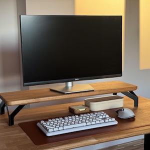 May include: A wooden monitor stand with black metal legs supporting a black Dell computer monitor. Below the stand sits a white mechanical keyboard, a grey computer mouse, a small wooden box, and a rectangular cream-colored container on a brown desk mat.