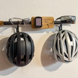 Può includere: Un supporto a parete con una barra orizzontale nera e un blocco centrale in legno inciso con il testo DISCIPLINE EQUALS FREEDOM. Il supporto sostiene due caschi da bicicletta, uno nero opaco e uno argento, oltre a due paia di occhiali da ciclismo.