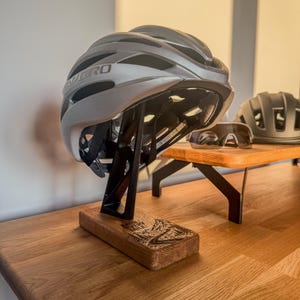 Könnte beinhalten: Ein silberner Giro-Fahrradhelm auf einem schwarzen Ständer mit einem Holzsockel, der eine eingravierte Berggrafik zeigt. Eine Radsportbrille und ein zweiter mattschwarzer Helm stehen auf einem Holzregal im Hintergrund.