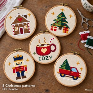 May include: Five circular wooden embroidery hoops containing cross-stitch designs on beige fabric. The patterns feature a gingerbread house, a Christmas tree with gifts, a red mug with the text COZY, a nutcracker, and a red truck carrying a tree.