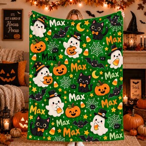 May include: A green fleece blanket featuring a repeating Halloween pattern of white ghosts wearing hats, black cats with orange bows, orange pumpkins, bats, spiderwebs, yellow stars, candy corn, and the name Max printed in orange text.