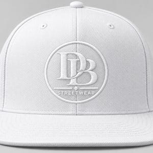 Op de afbeelding: Een effen witte honkbalpet met een platte klep en een gestructureerde kroon. In het midden aan de voorzijde bevindt zich een geborduurd cirkelvormig logo met de letters DB en het woord STREETWEAR in wit garen voor een monochroom ontwerp.