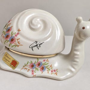 Puede incluir: Una figura de caracol de cerámica blanca con acabado brillante, que presenta una concha en espiral decorada con motivos florales rosas y azules. La concha tiene una firma negra y un borde dorado. Una pegatina dorada dice Esclusiva Creazione Made in Italy.