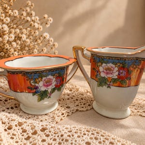Puede incluir: Un juego de azucarero y lechera de porcelana con base blanca y acentos vibrantes en naranja, azul y dorado. Las piezas están decoradas con coloridos motivos florales, bordes festoneados y asas con ribetes dorados, mostrando un diseño clásico.