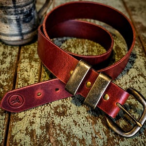 May include: A deep red leather belt featuring a distressed finish, two wide brass-toned metal loops, and a matching brass-toned buckle. The pointed end of the belt displays a circular embossed logo with the letter T inside a circle.