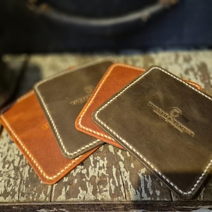 May include: A set of square leather coasters in shades of brown and dark grey, featuring rounded corners, white contrast stitching along the edges, and the embossed logo text Thread & Temper with a circular icon in the centre of each piece.